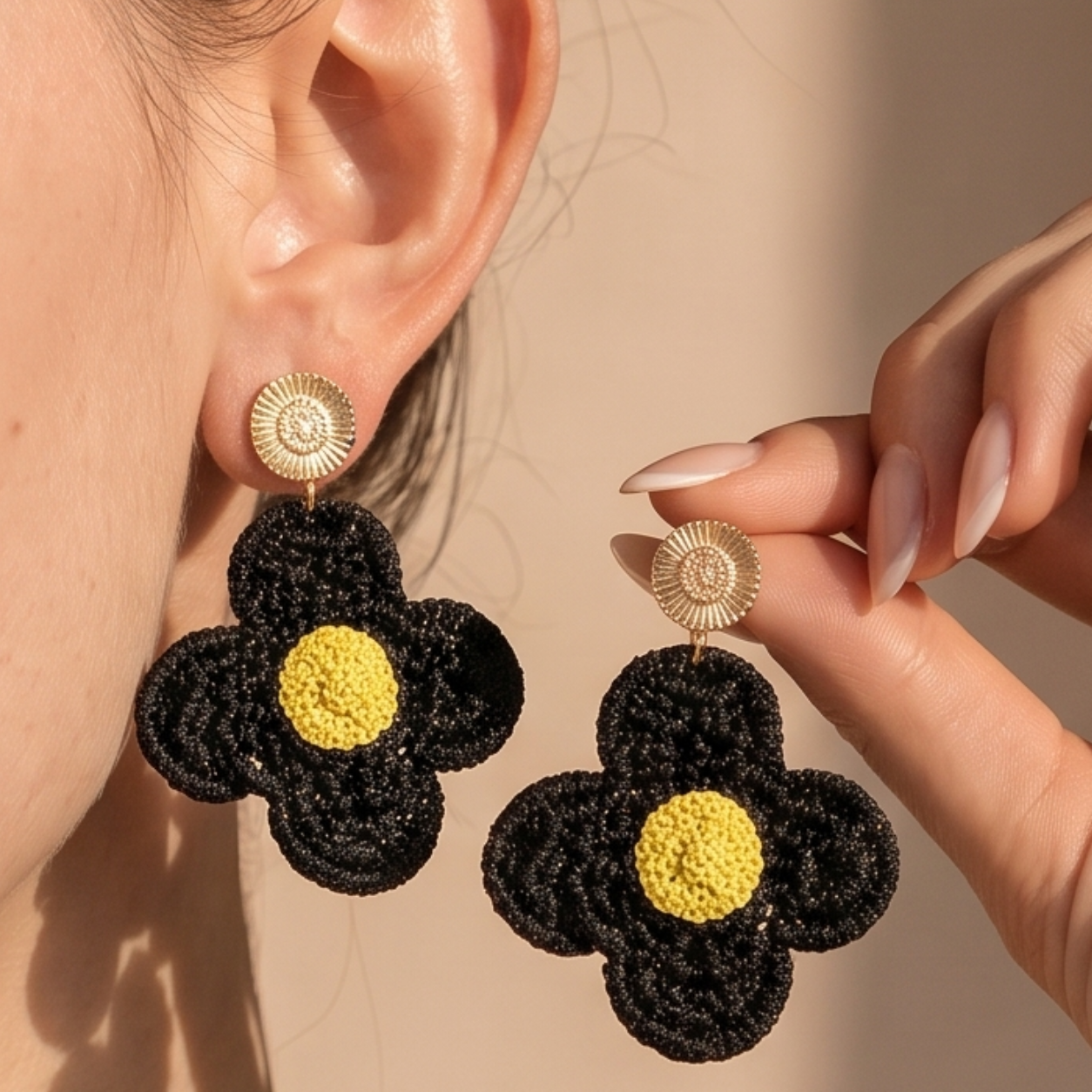 Image of Black Boho Crochet Sunflower Earrings, shown worn on an ear and held by a hand against a warm beige background.