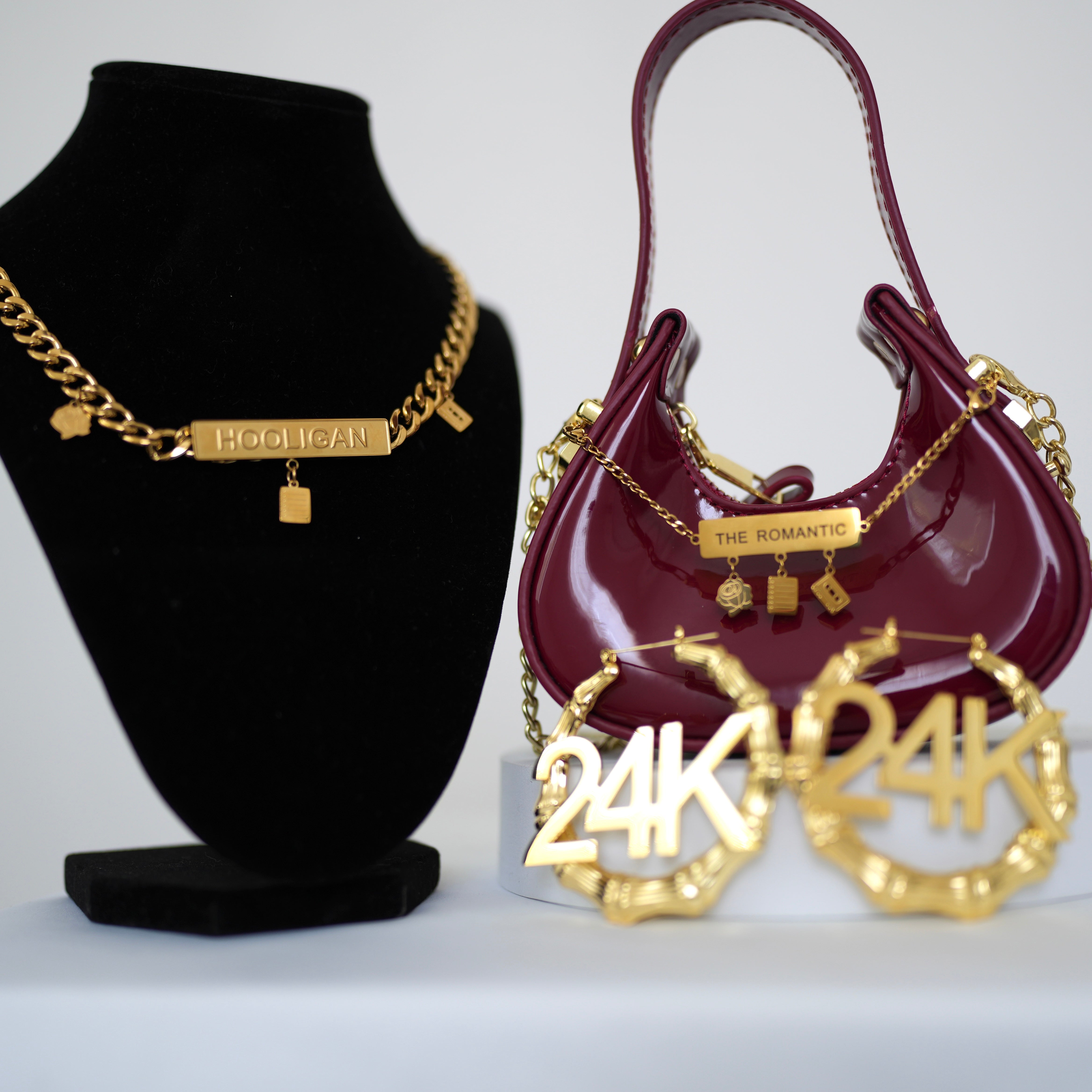 Image of Bruno Mars Hooligan Gold necklace, maroon handbag with gold chain, and gold '24K' earrings on a white background