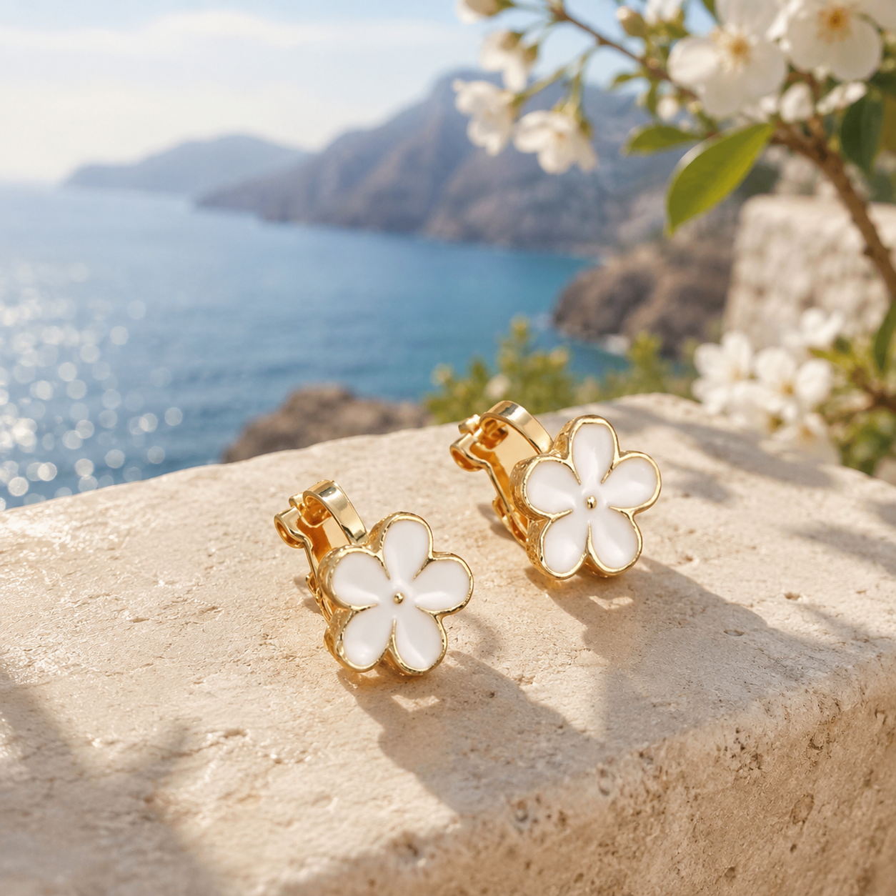 Image of A pair of white and gold flower-shaped clip-on earrings with white enamel centers, resting on a stone ledge with a scenic blue sea and mountains in the background.