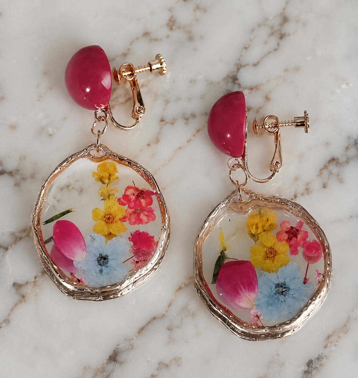Image of A pair of rose gold clip-on earrings with a dome of preserved flowers and petals, resting on a white marble surface.