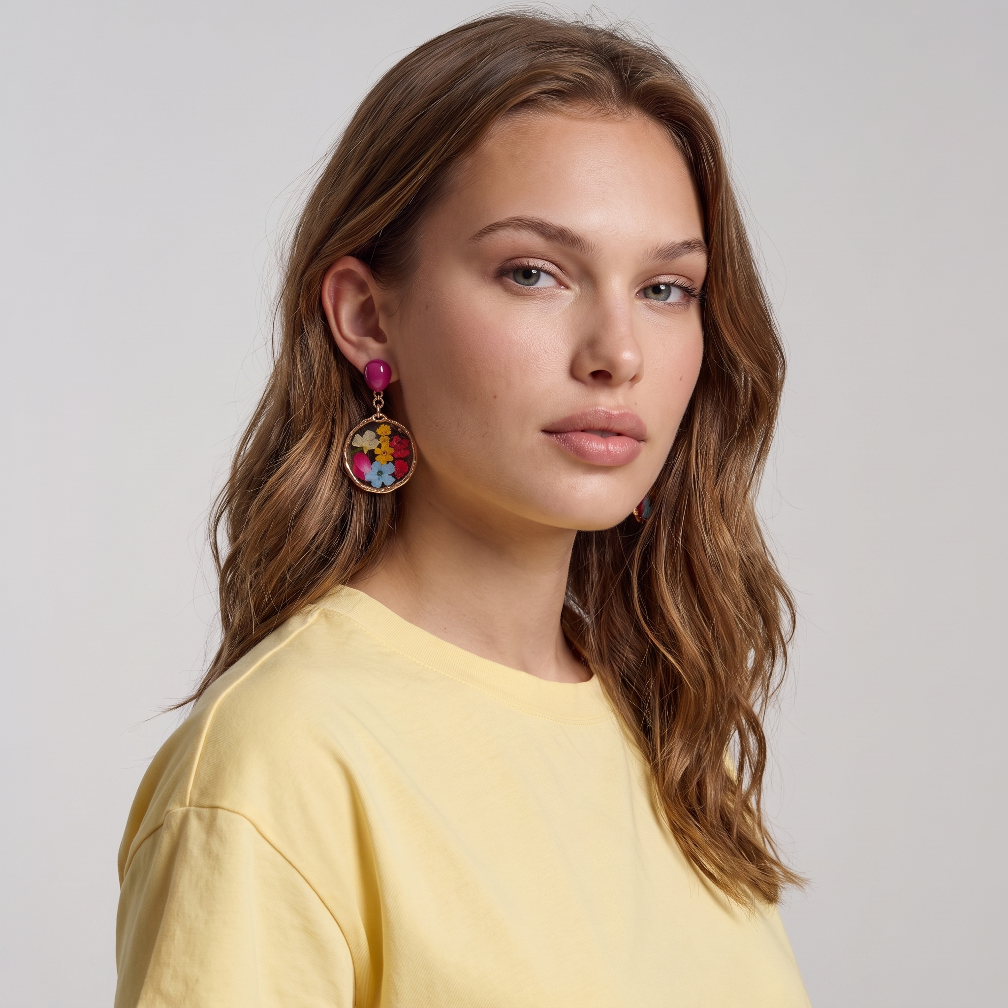 Image of A woman with wavy light brown hair wearing a pale yellow t-shirt and a colorful dangling clip on earring, photographed against a neutral light gray background.