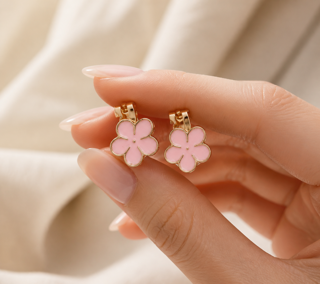 Image of A close-up of a hand holding a pair of gold-tone flower-shaped clip on earrings with light pink enamel centers, set against a soft beige fabric background.