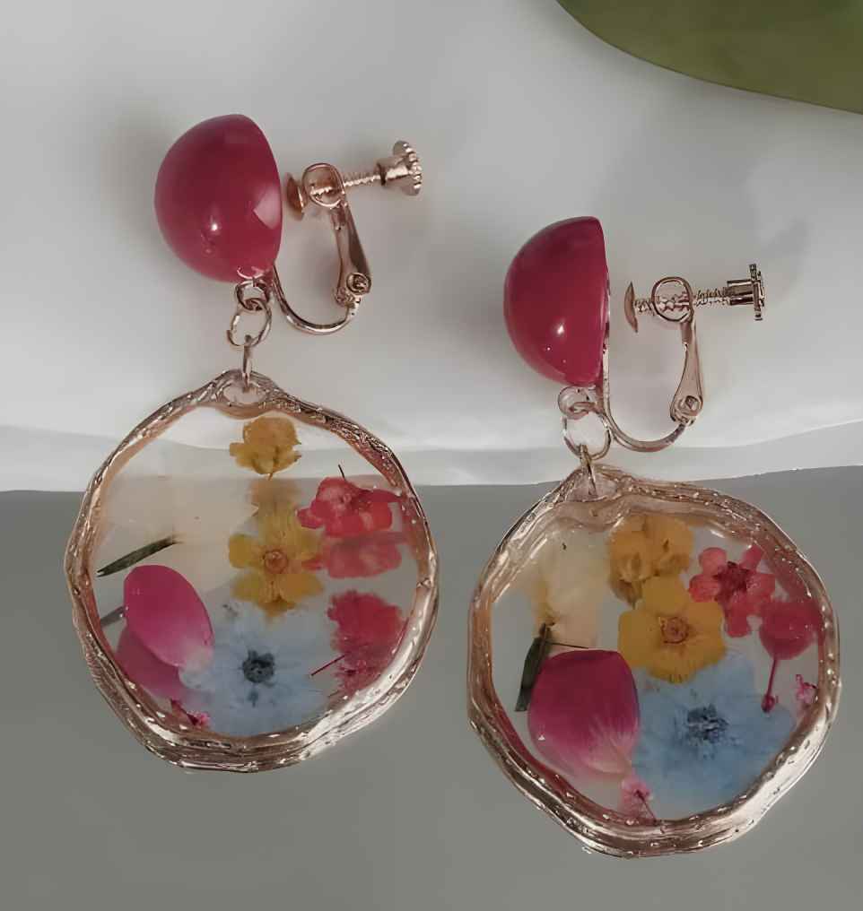 Image of A pair of rose gold clip on earrings with large, irregular clear resin pendants containing pressed flowers in pink, yellow, and blue, topped with glossy dark pink dome beads.