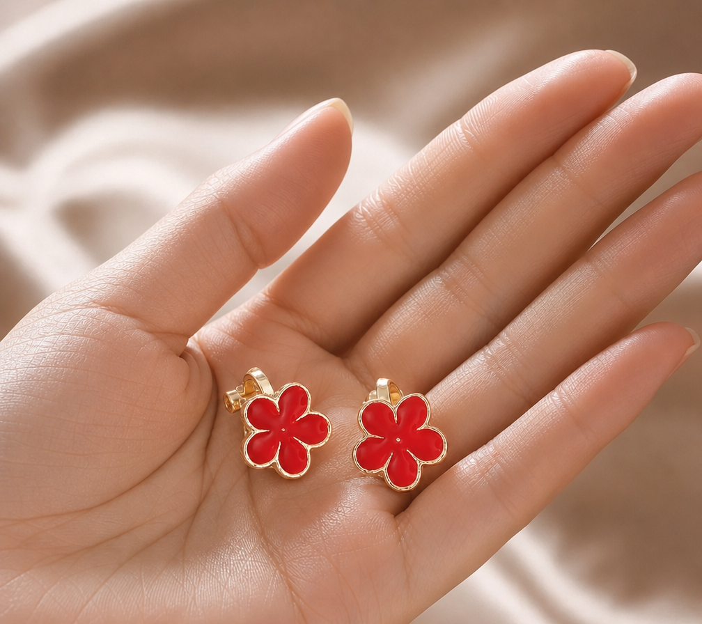 Image of A pair of gold-tone flower-shaped  clip on earrings with bright red enamel centers, held in a person's palm against a soft beige fabric background.