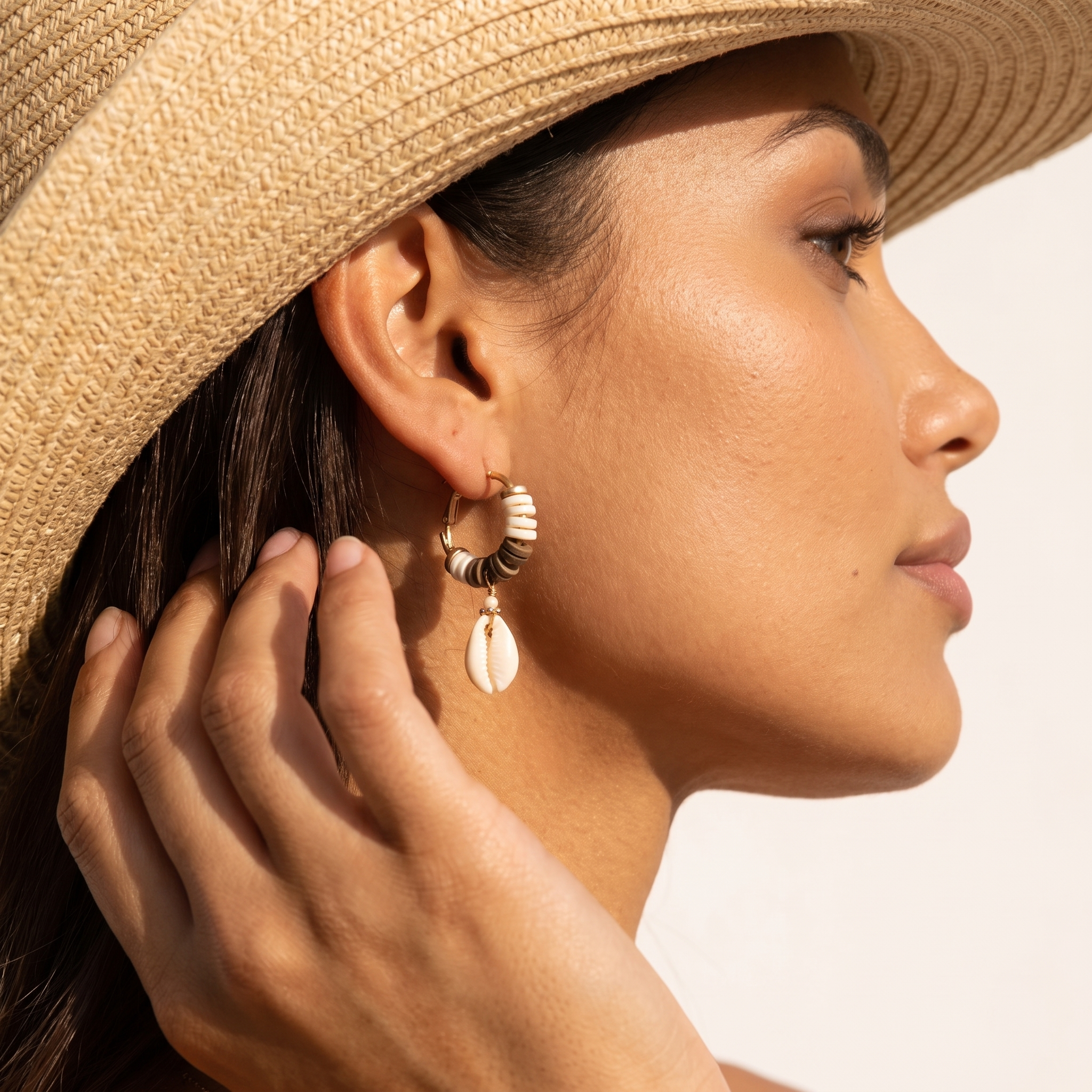 Image of Close-up of a woman wearing gold seashell earrings with a wood bead hoop and cowrie shell drop, styled with a straw cowgirl hat