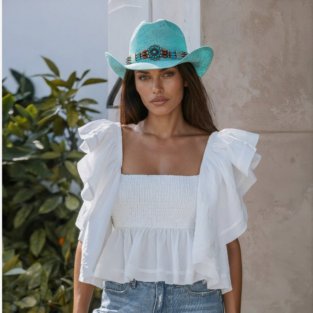 Image of A woman wearing a turquoise cowboy hat with a beaded band and a white smocked ruffle-sleeve top, paired with light blue jeans, standing against a neutral wall with green foliage on the side.
