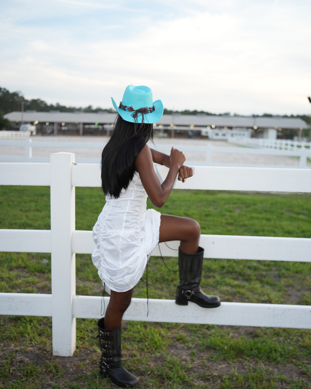 Image of A woman wearing a white ruffled dress, a bright turquoise cowboy hat, and black boots sits on a white fence in an outdoor paddock with a horse fence and stable buildings in the background.