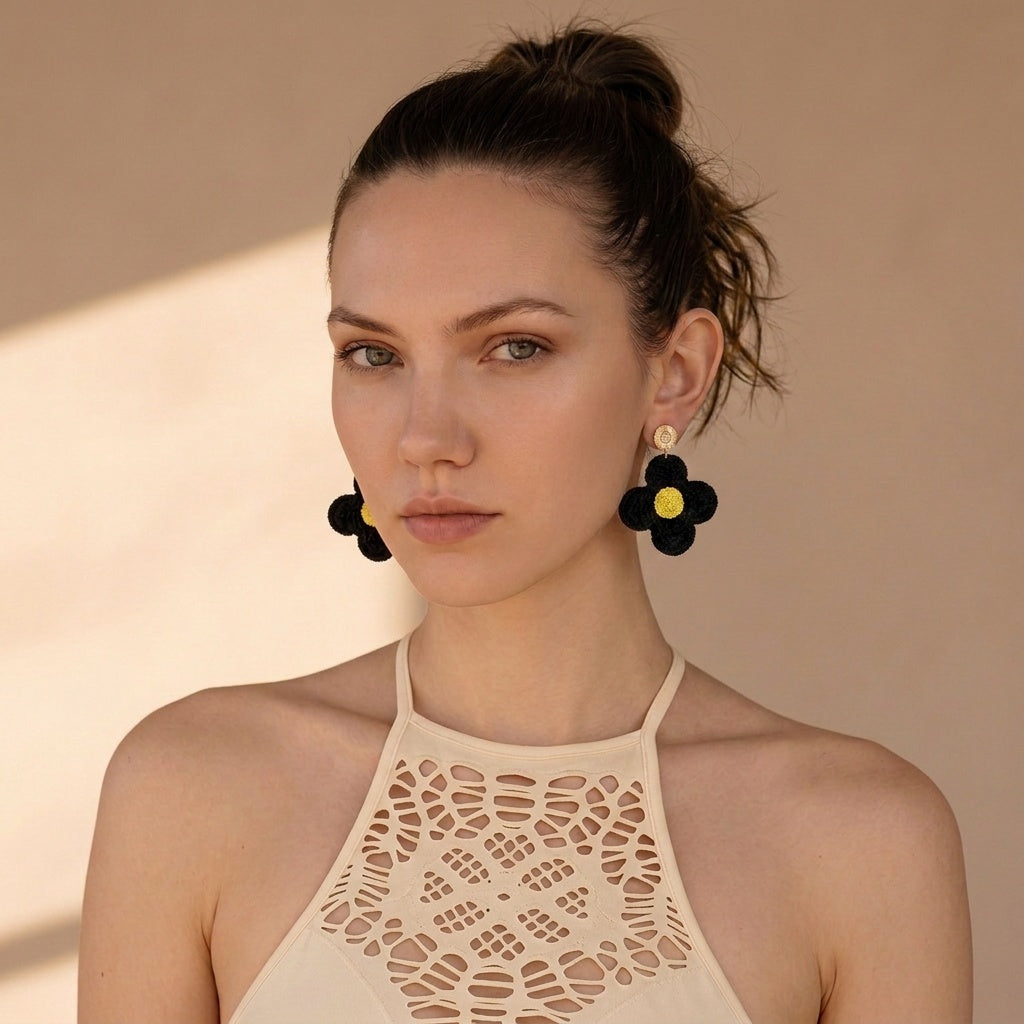 Image of A woman wearing black crochet flower-shaped dangle earrings with bright yellow centers, paired with a beige halter top against a warm beige background.