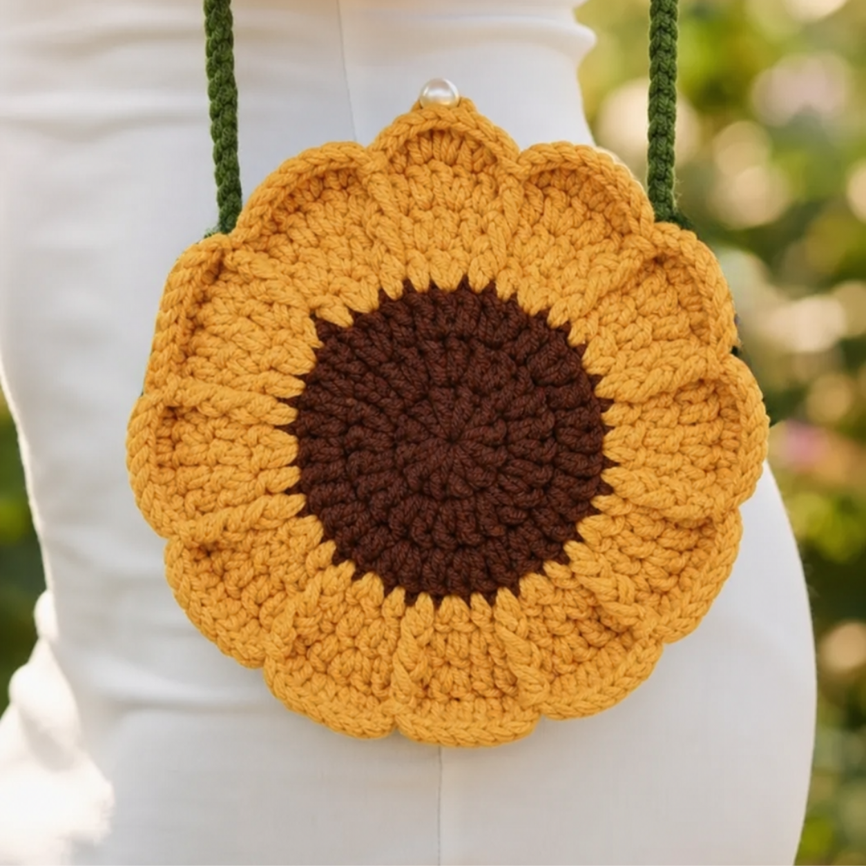Image of A crocheted sunflower purse with a yellow and brown flower face and green shoulder straps, worn on a person's hip against a blurred green outdoor background.