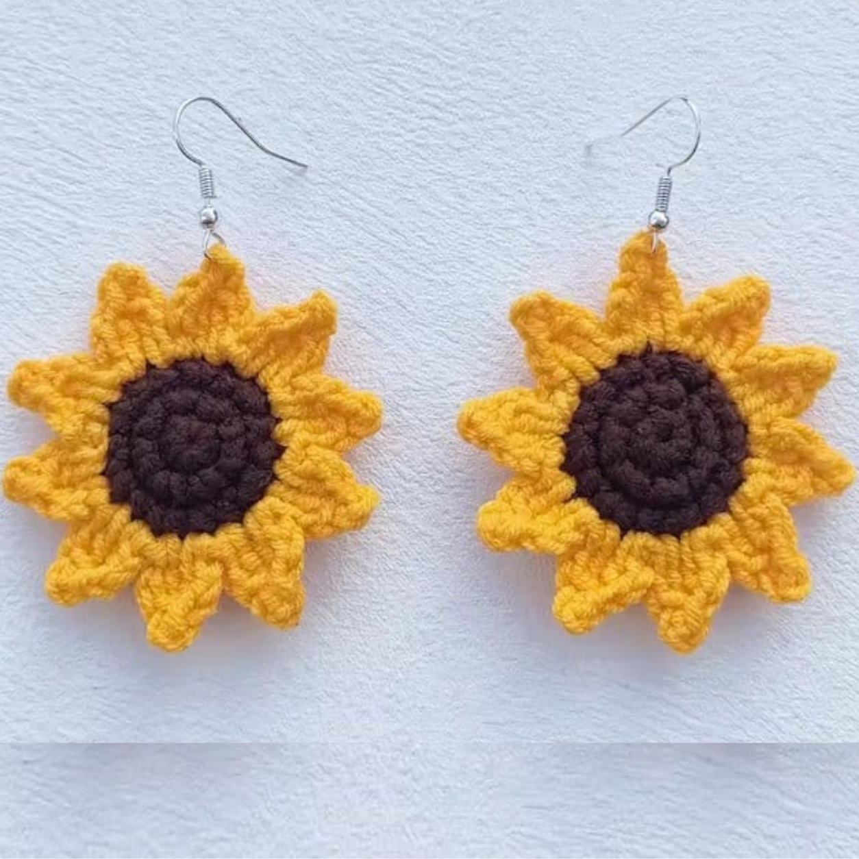 Image of A pair of handmade crocheted sunflower dangle earrings with yellow petals, dark brown centers, and silver fishhook wires, displayed on a textured white background.