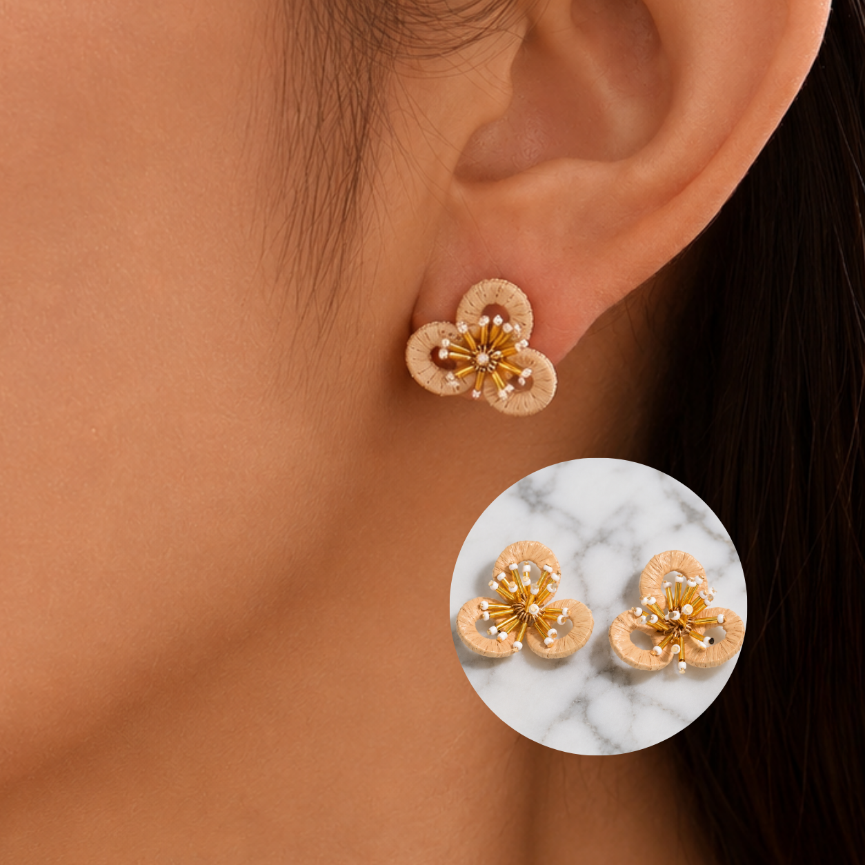Image of A close-up of a person's ear wearing a small raffia flower-shaped stud earring with beige petals and a gold center accented by tiny white pearls. A circular inset shows two matching earrings on a white marble background.