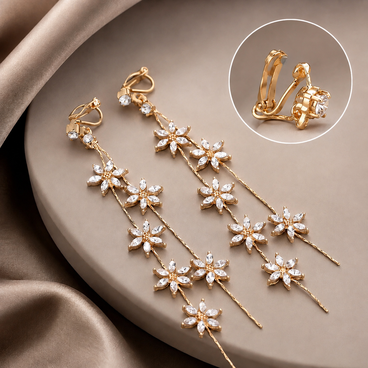 Image of A pair of gold dangle clip on earrings with floral clusters of clear rhinestones, displayed on a beige satin surface with a circular inset showing the hinged clasp detail.
