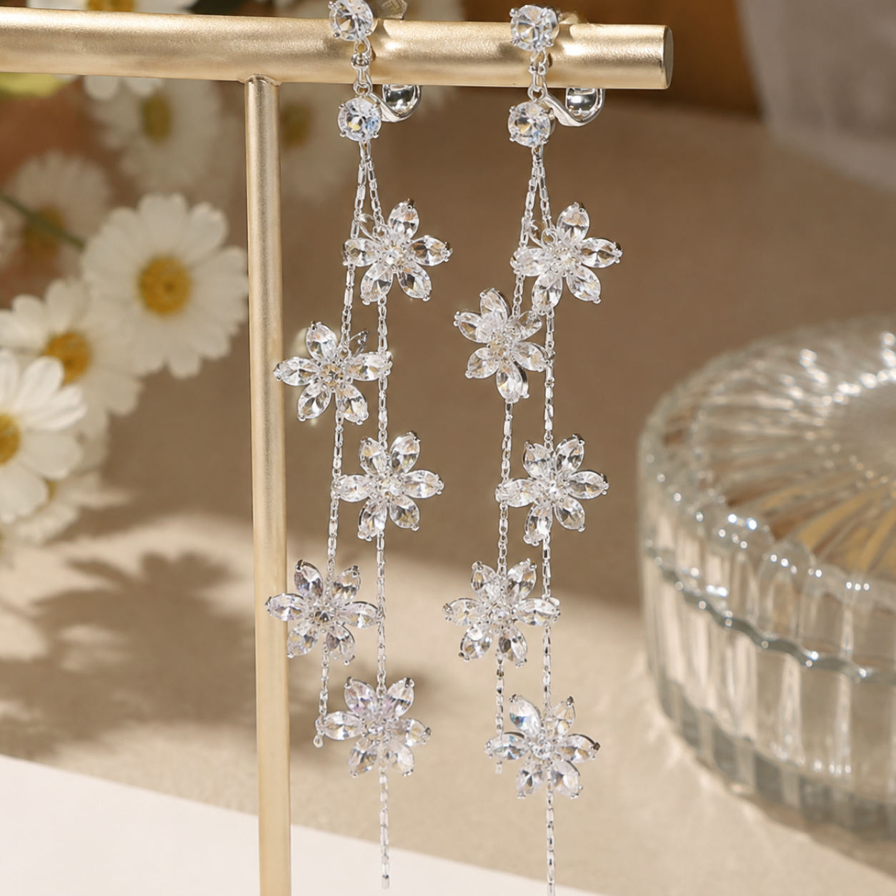 Image of A pair clip on earrings featuring long dangling crystal flowers hanging from a gold display stand, with soft-focus flowers and a glass dish in the background.