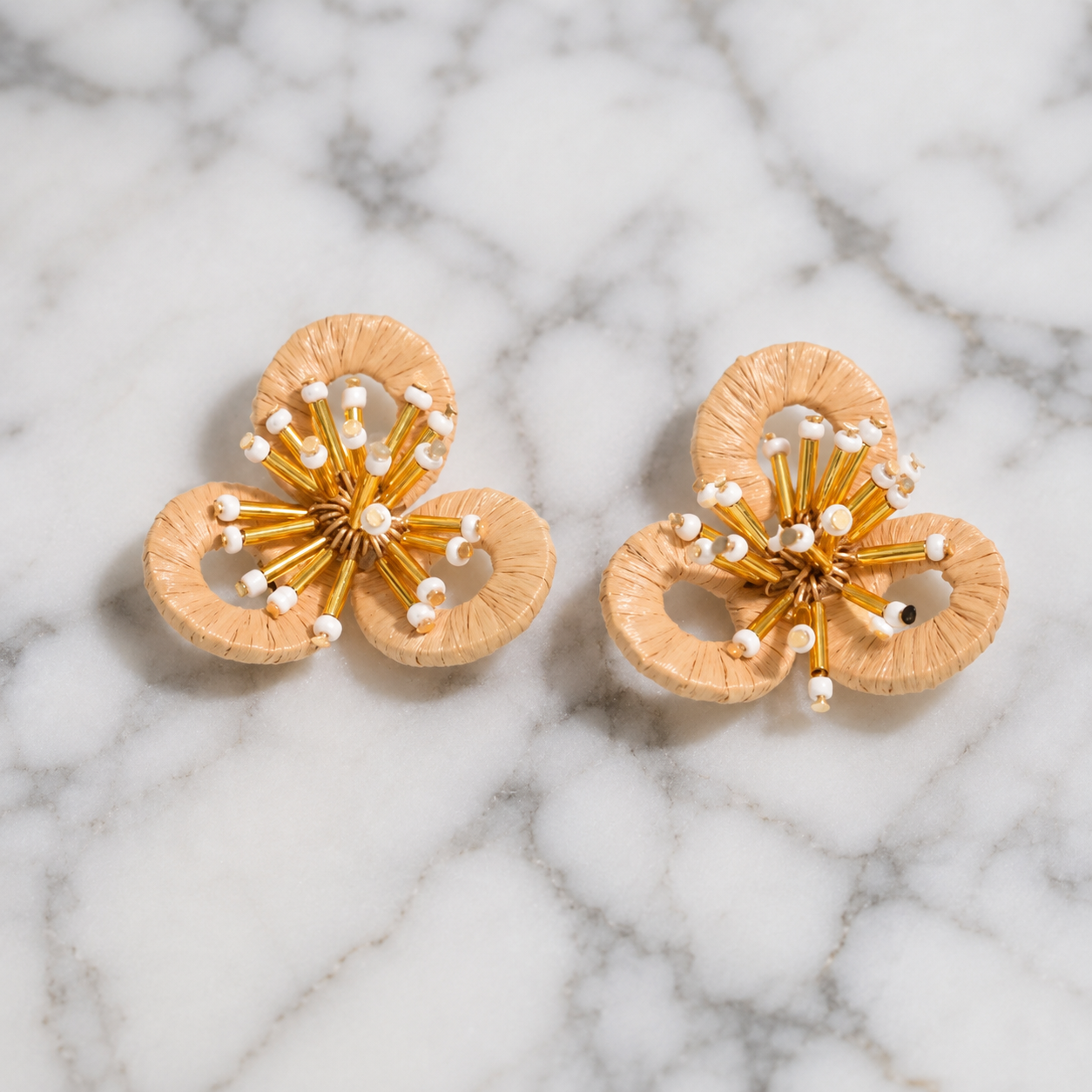 Image of A pair of handmade floral earrings with light wood petals and gold wire centers, accented with small white beads, displayed on a white and gray marble surface.