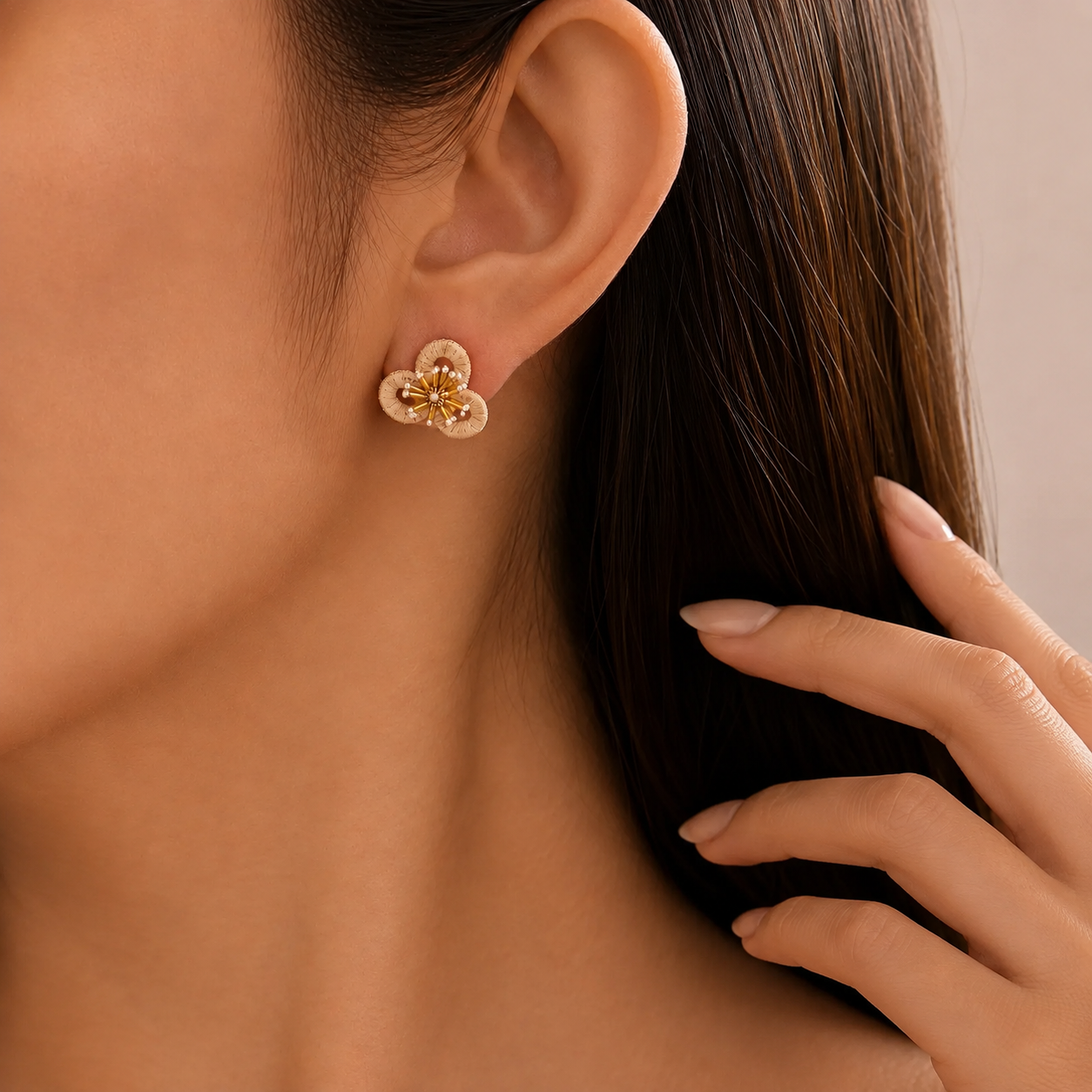 Image of A close-up of a woman's ear wearing a small straw flower-shaped stud earring with gold-toned petals and a pale pink center, against a soft beige background.