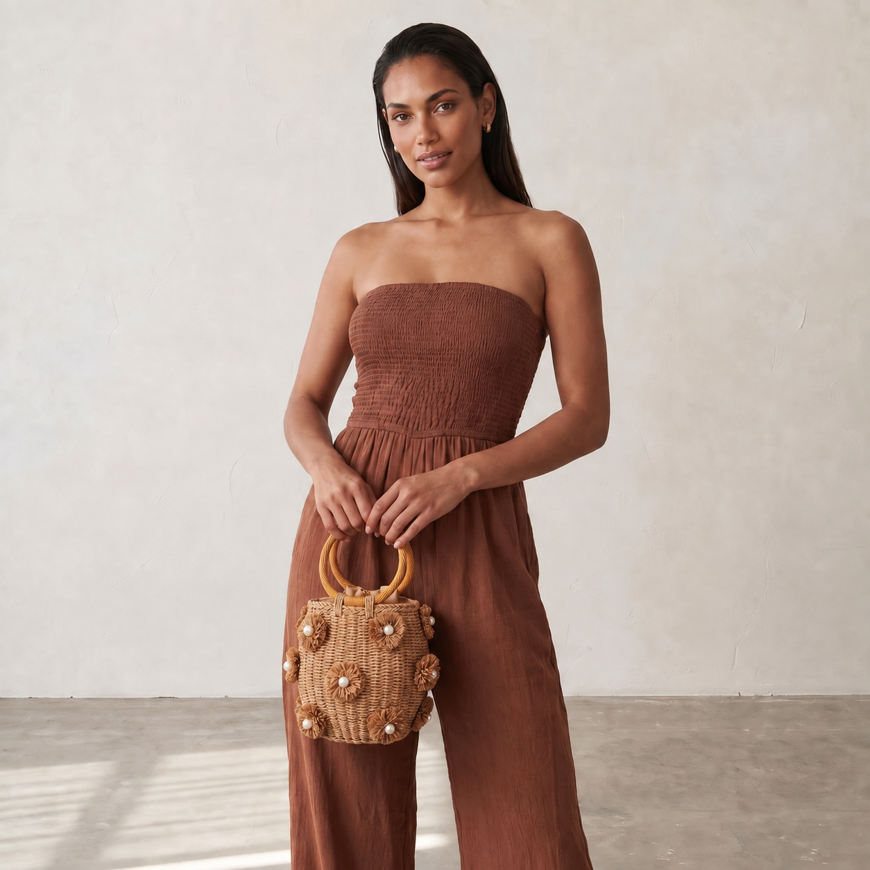 Image of A woman stands indoors holding a woven tan handbag with round wooden handles and pearl-like embellishments, against a plain light wall.