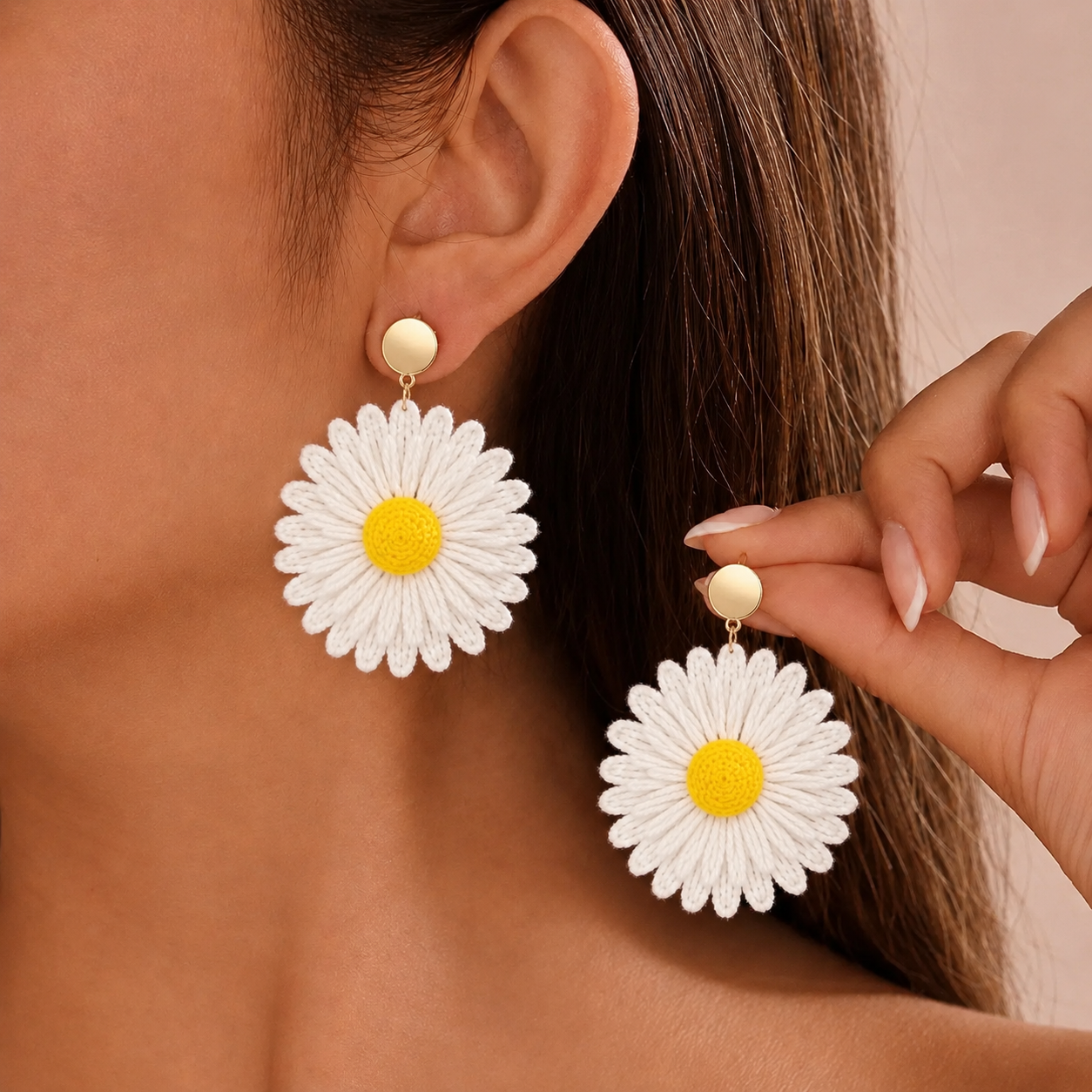 Image of A close-up lifestyle shot of a model wearing a pair of boho daisy drop earrings with yellow centers and gold-toned studs. The model's ear, side profile, and hand holding one of the earrings are visible against a soft beige background.