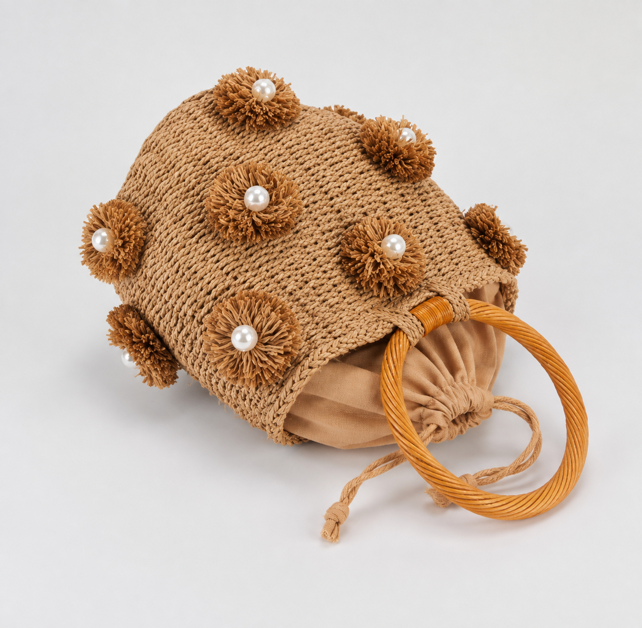 Image of A tan woven sun hat with decorative pom-pom flowers and pearl accents, featuring a drawstring closure and a thick braided straw handle, photographed on a light gray background.