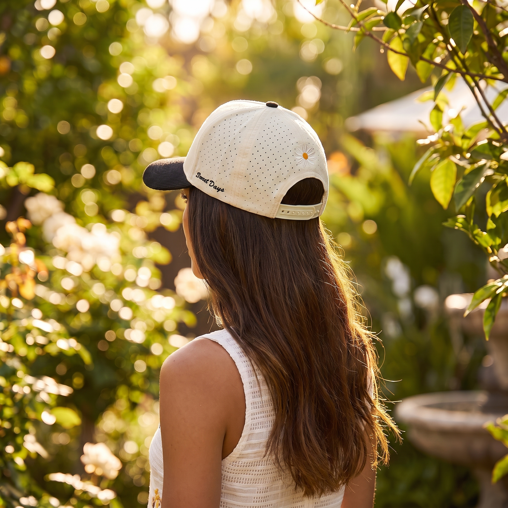 Image of Back view of a retro trucker hat showing the embroidered daisy detail on the crown, woman in a garden setting