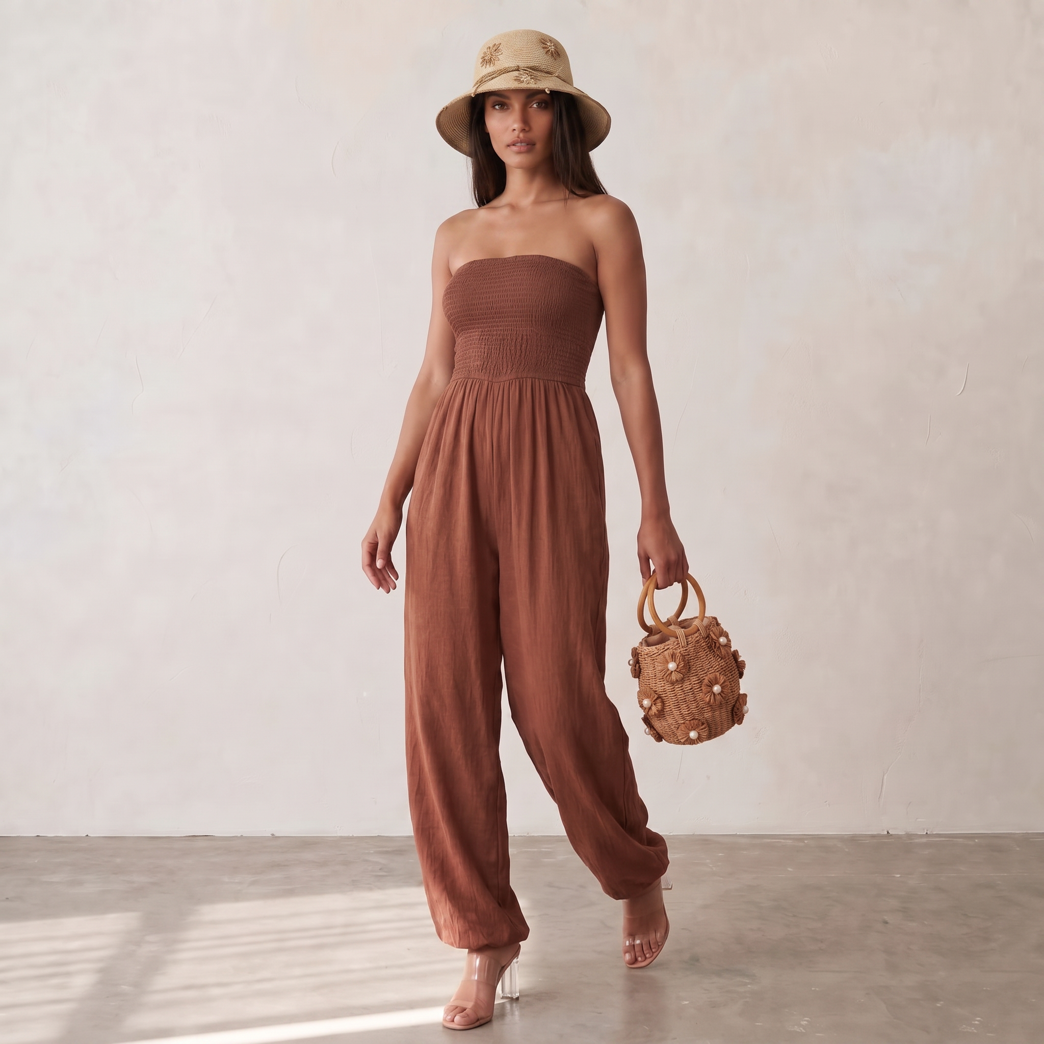 Image of A woman stands in a studio wearing a strapless rust-brown jumpsuit with wide legs, a beige sun hat with floral embroidery, and holding a small woven brown handbag.