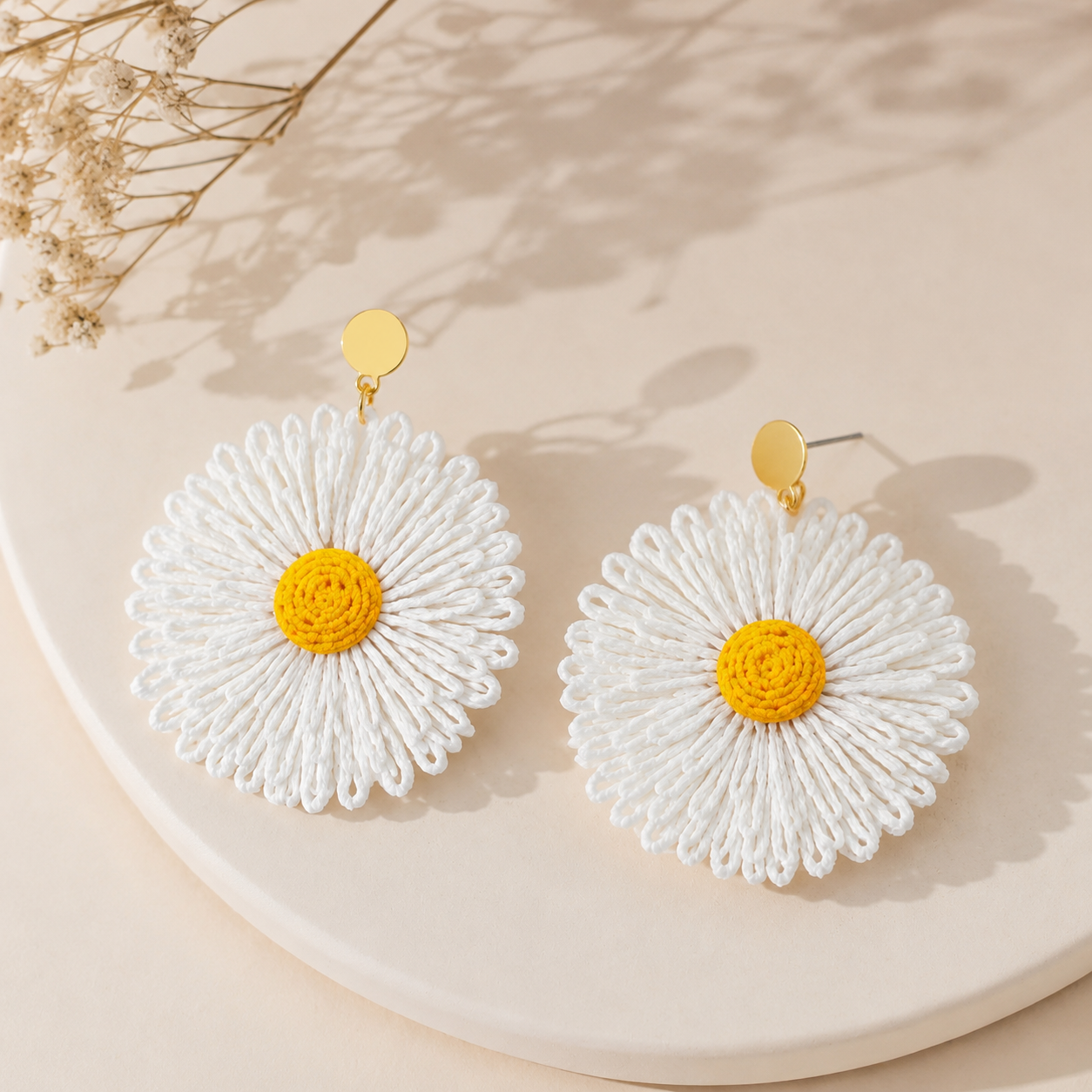 Image of A pair of large flower-shaped daisy earrings with white raffia petals, bright yellow centers, and textured gold posts, displayed on a neutral beige background.