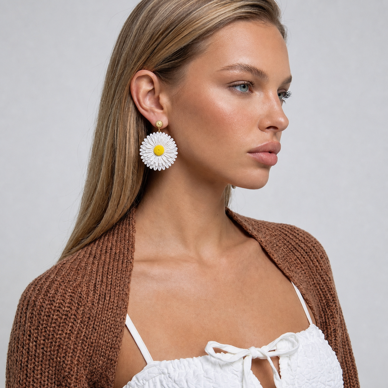 Image of A woman in profile wearing a large white flower-shaped dangle earring with a yellow center and a brown knit cardigan over a white top, against a light gray background.