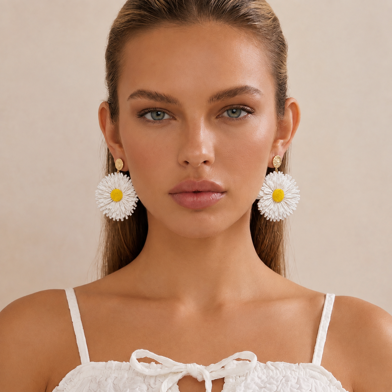Image of A close-up of a woman wearing large white straw flower earrings with yellow centers and gold-toned studs, paired with a white lace top against a soft beige background.