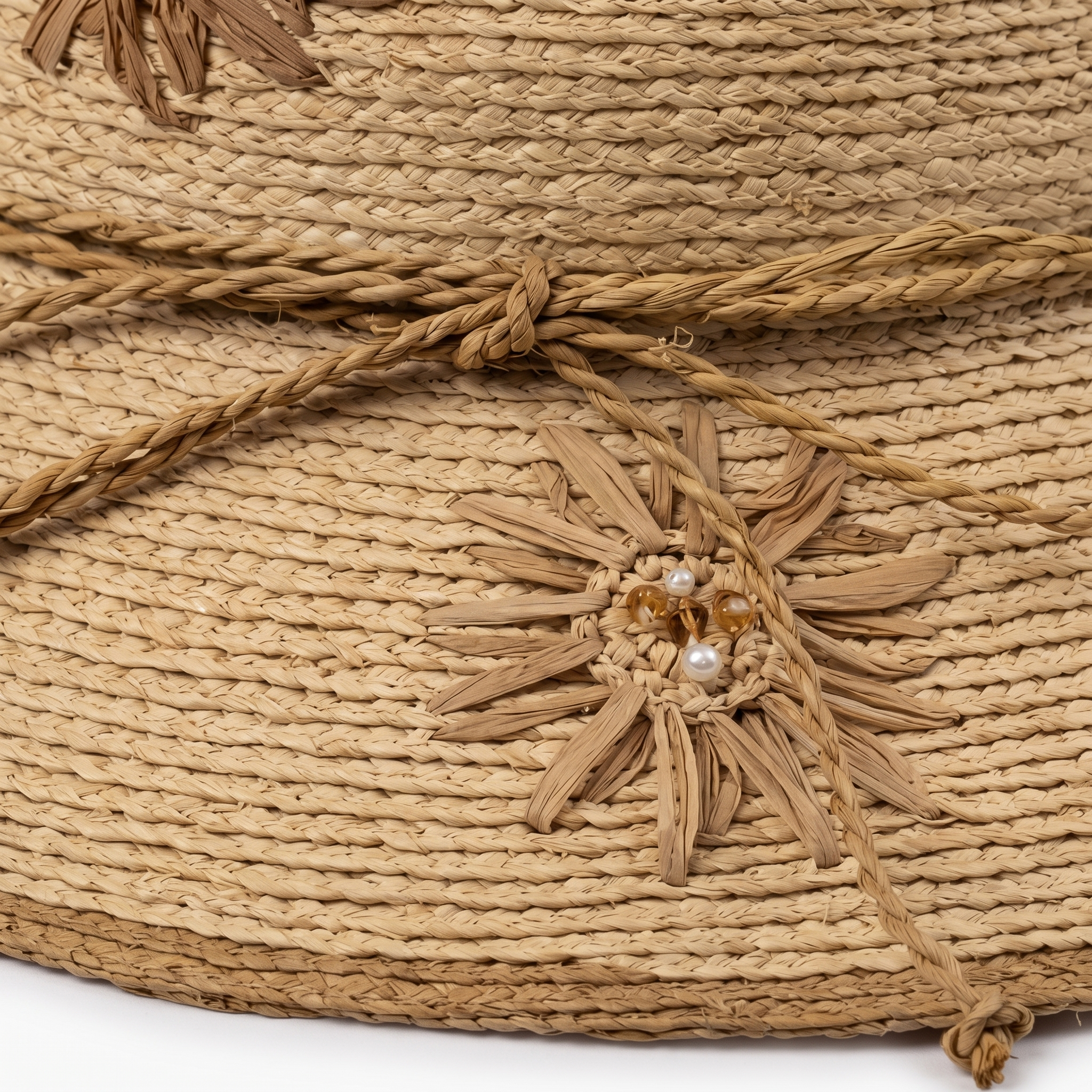 Image of Close-up of a natural straw hat with a braided rope band tied in a bow and a decorative woven flower embellishment with pearl-like beads.