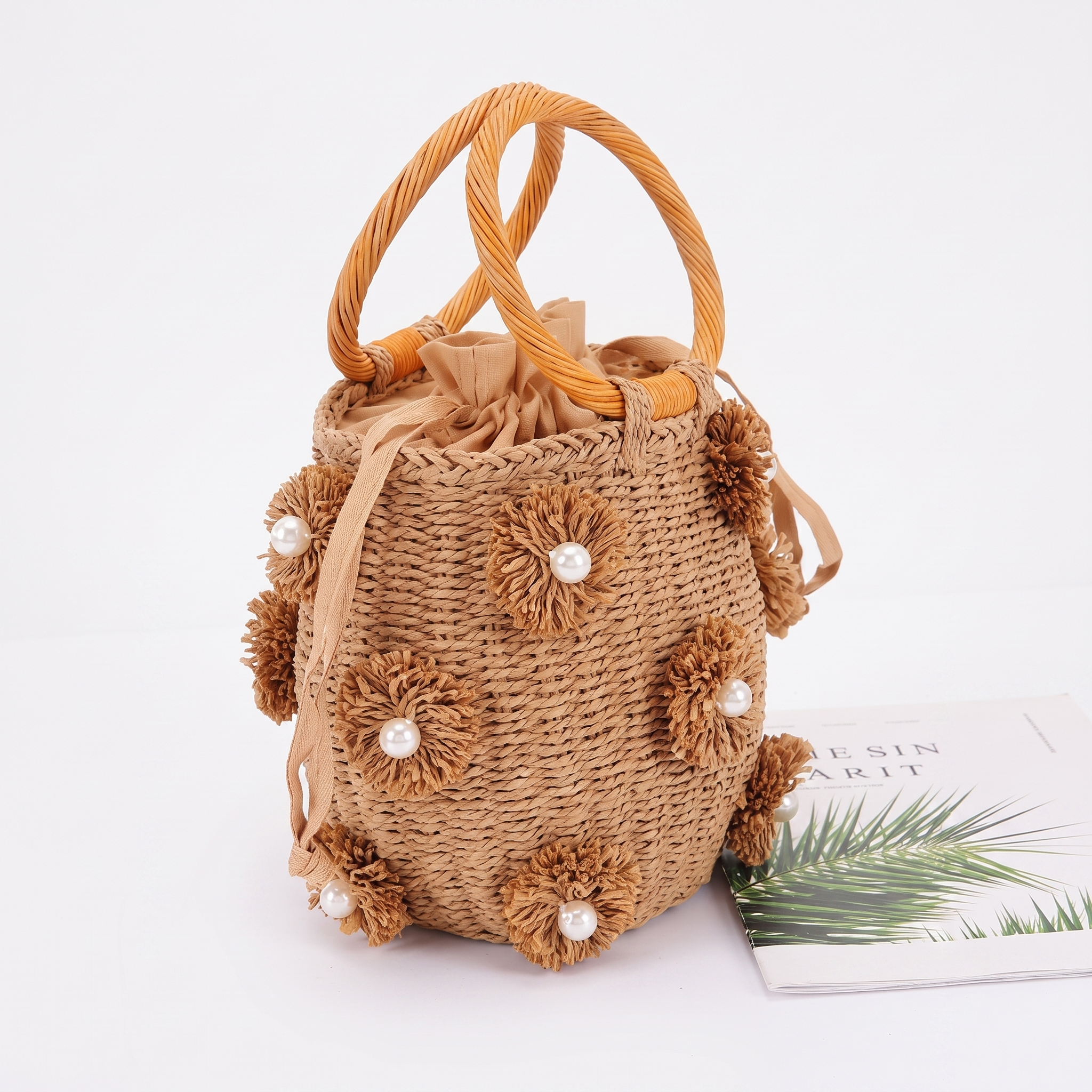 Image of A natural woven straw bucket bag with twisted rope handles, adorned with 3D braided flower embellishments and small white pearl accents. The bag is styled next to a magazine on a clean white background.