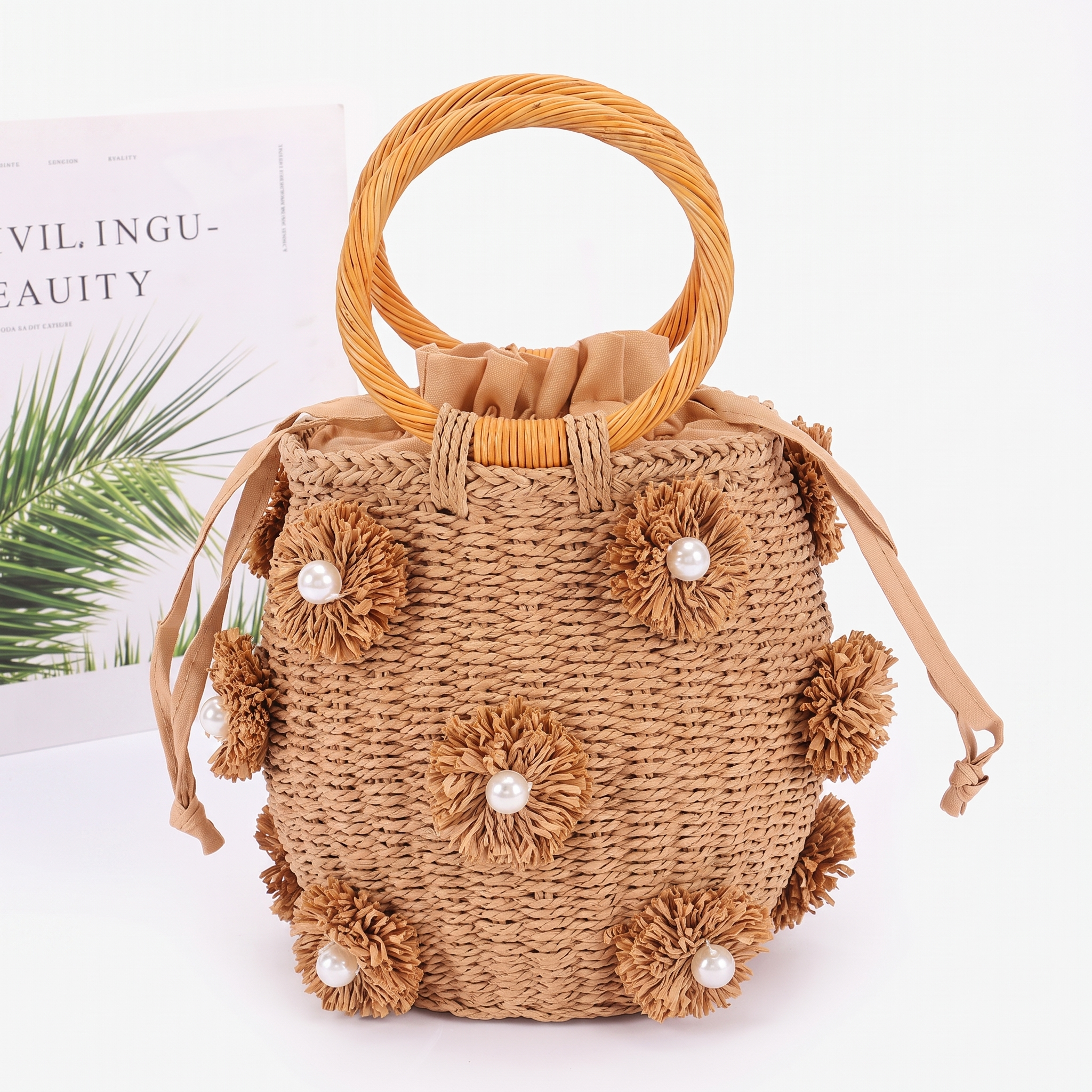 Image of A natural woven straw bucket bag with a round wooden handle, decorated with 3D frayed flower embellishments that have pearl-like centers, set against a clean white background.