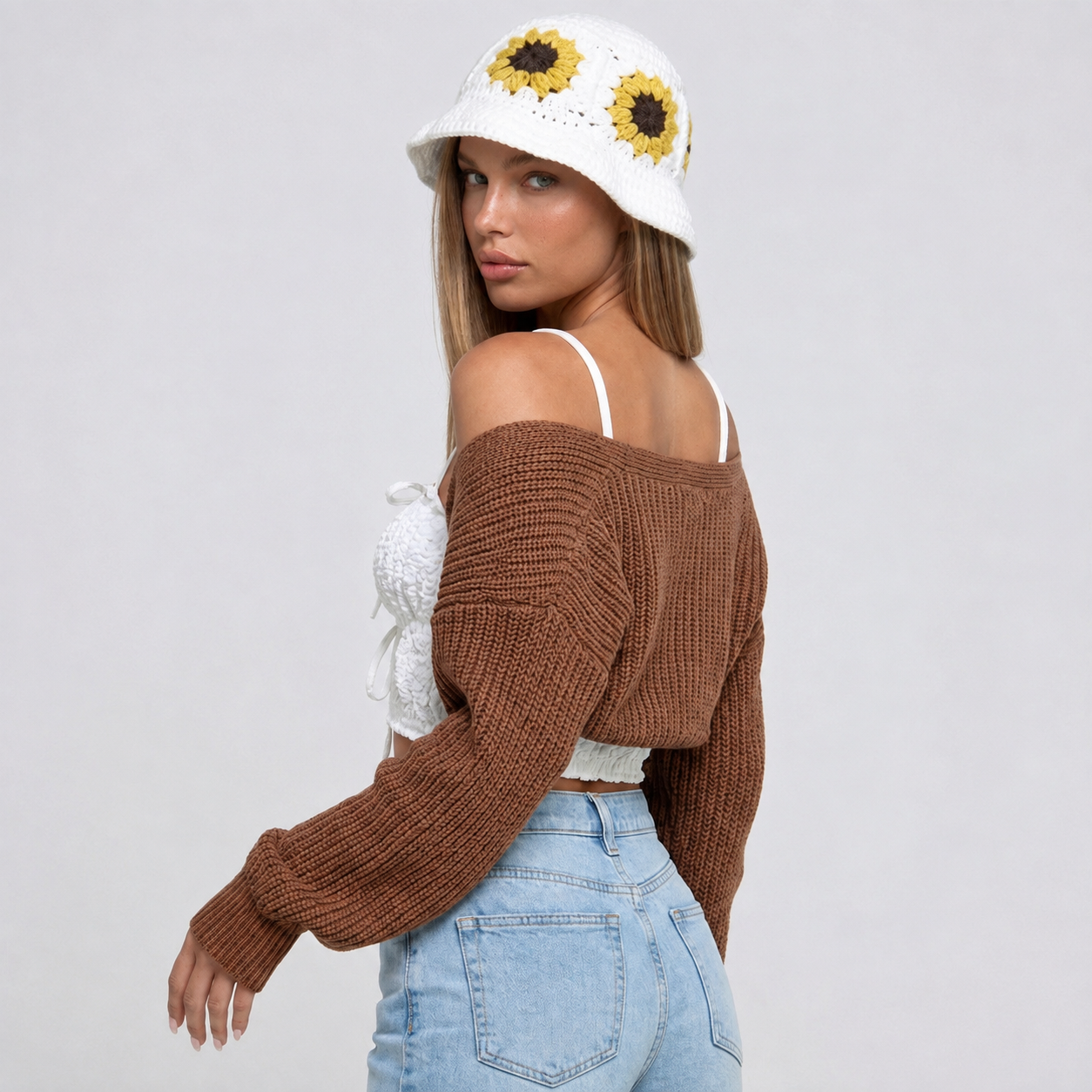Image of A woman wearing a brown chunky knit off-the-shoulder cropped sweater, light blue jeans, and a white crocheted bucket hat with yellow sunflower details, photographed against a light gray studio background from the rear.