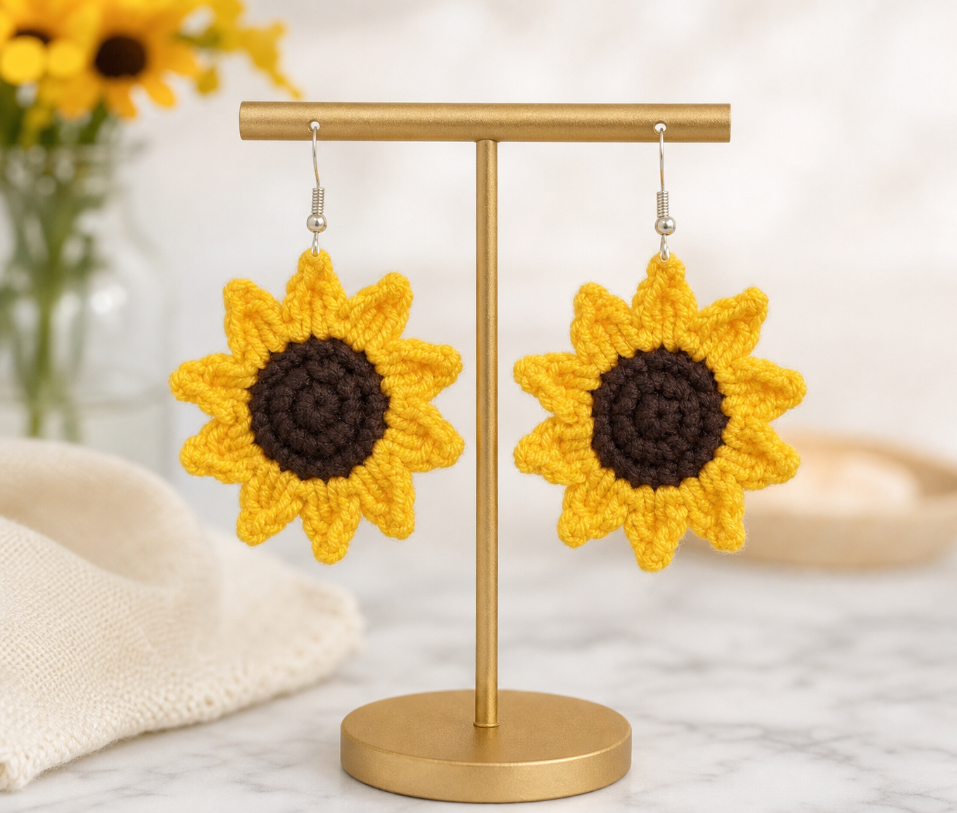 Image of Crocheted sunflower earrings displayed on a gold stand against a soft, blurred background with a marble surface and a vase of sunflowers.