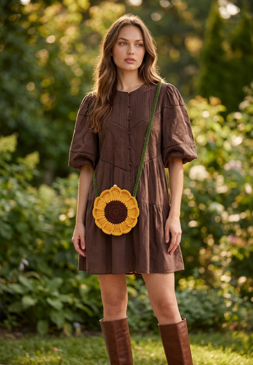 Image of A woman stands outdoors in a garden wearing a crocheted sunflower-shaped crossbody bag with a green strap.