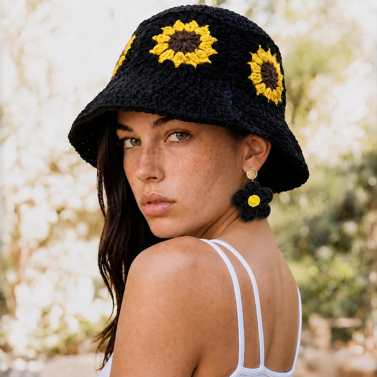 Image of A woman wearing a black crocheted bucket hat with yellow sunflower details, photographed outdoors with a soft blurred background.