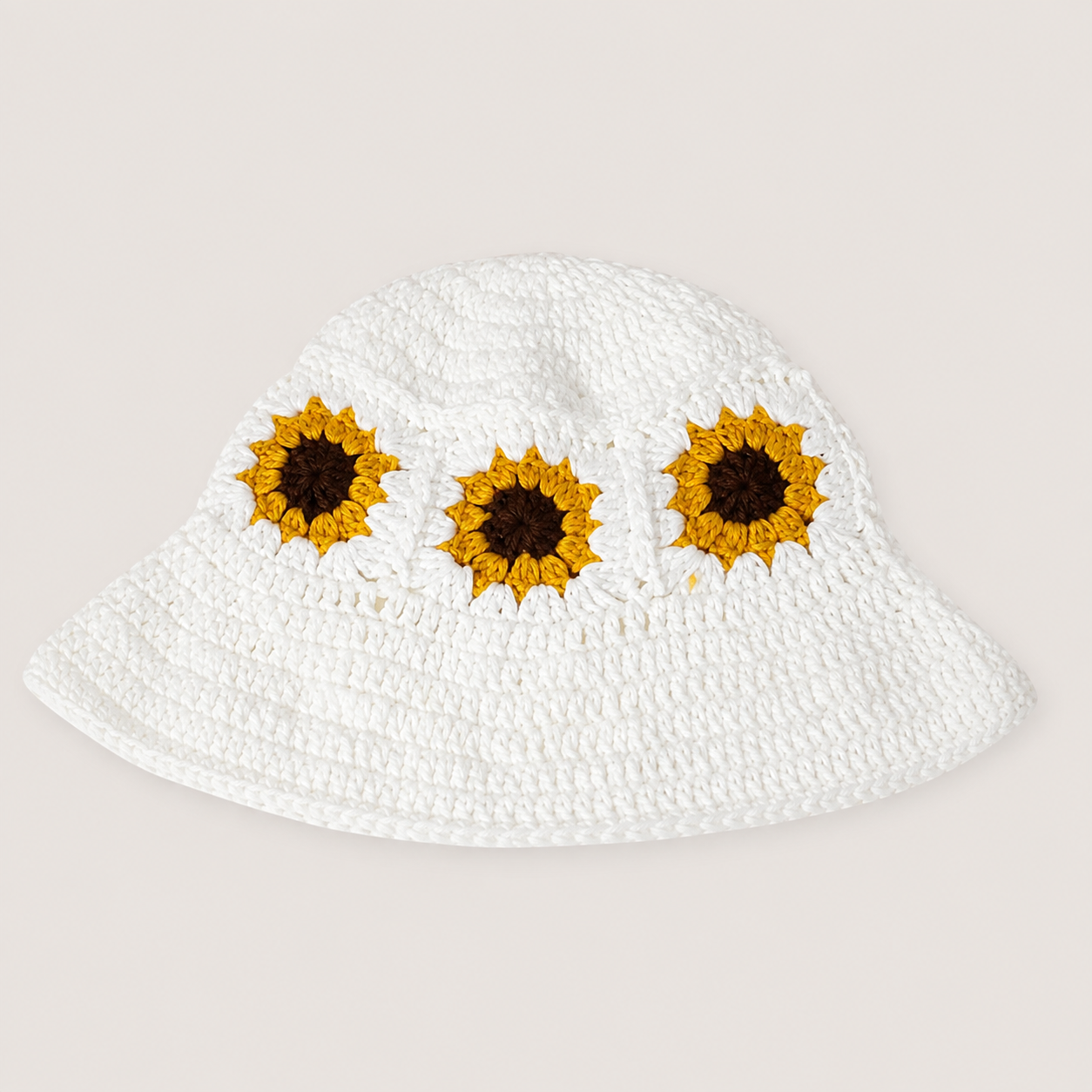 Image of Flatlay of a white crocheted bucket hat featuring three embroidered sunflower motifs across the front, displayed against a solid light beige background.