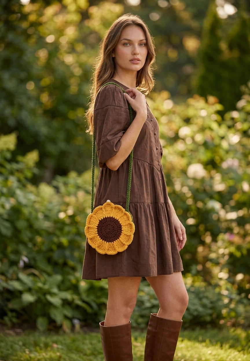 Image of Model standing outdoors in a garden carrying a crocheted sunflower-shaped crossbody bag with a green strap.