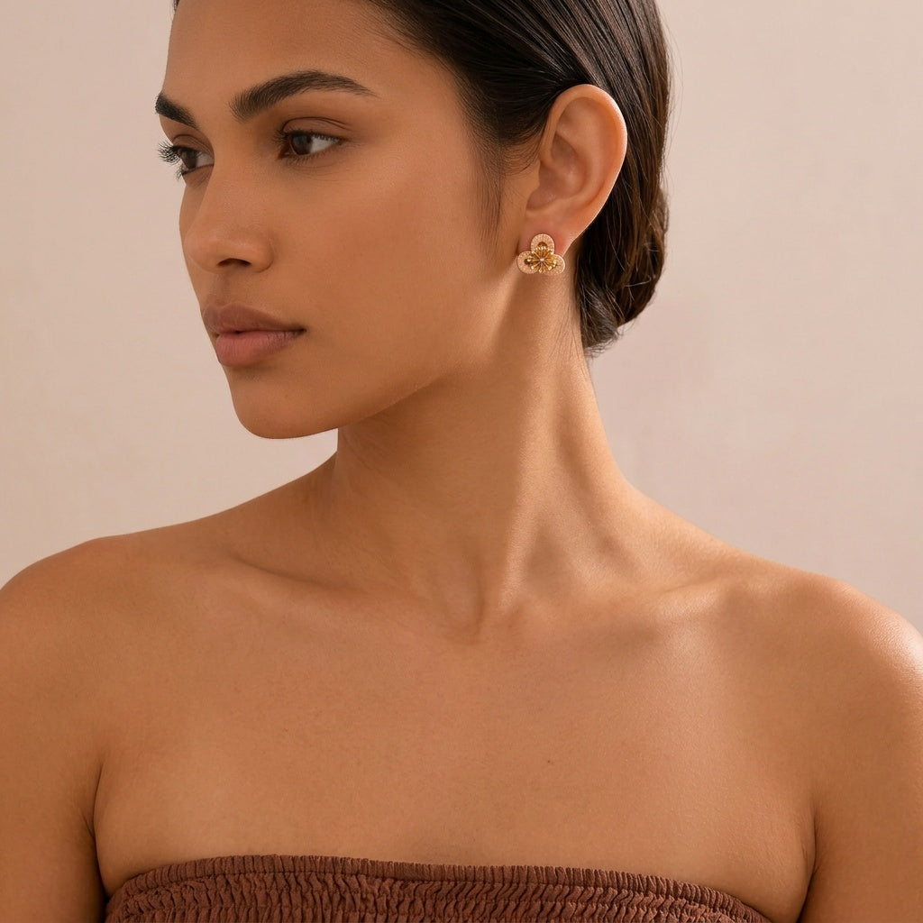 Image of A close-up of a woman wearing a small gold-toned floral stud earring, shown from the chest up against a soft beige background.