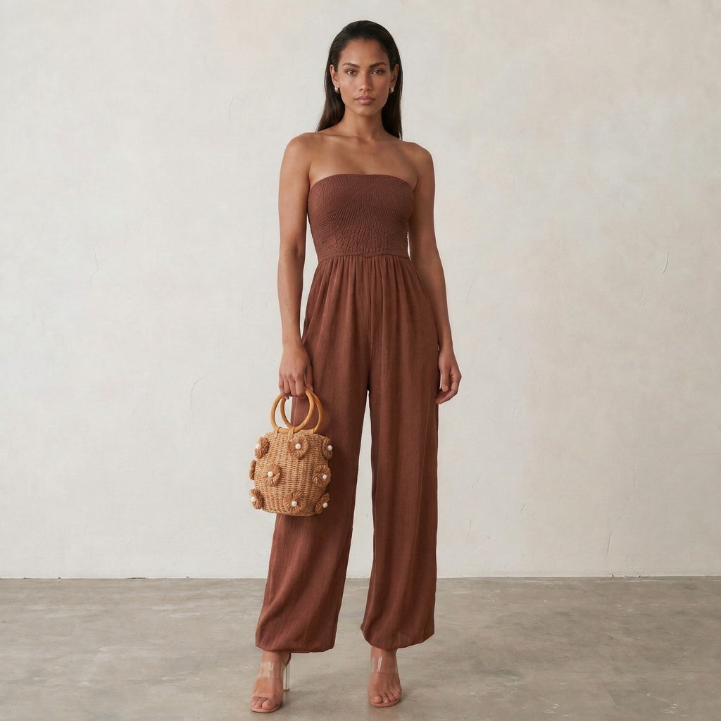 Image of A woman stands against a light beige textured wall wearing a strapless brown jumpsuit and holding a small woven tan handbag with wooden handles.